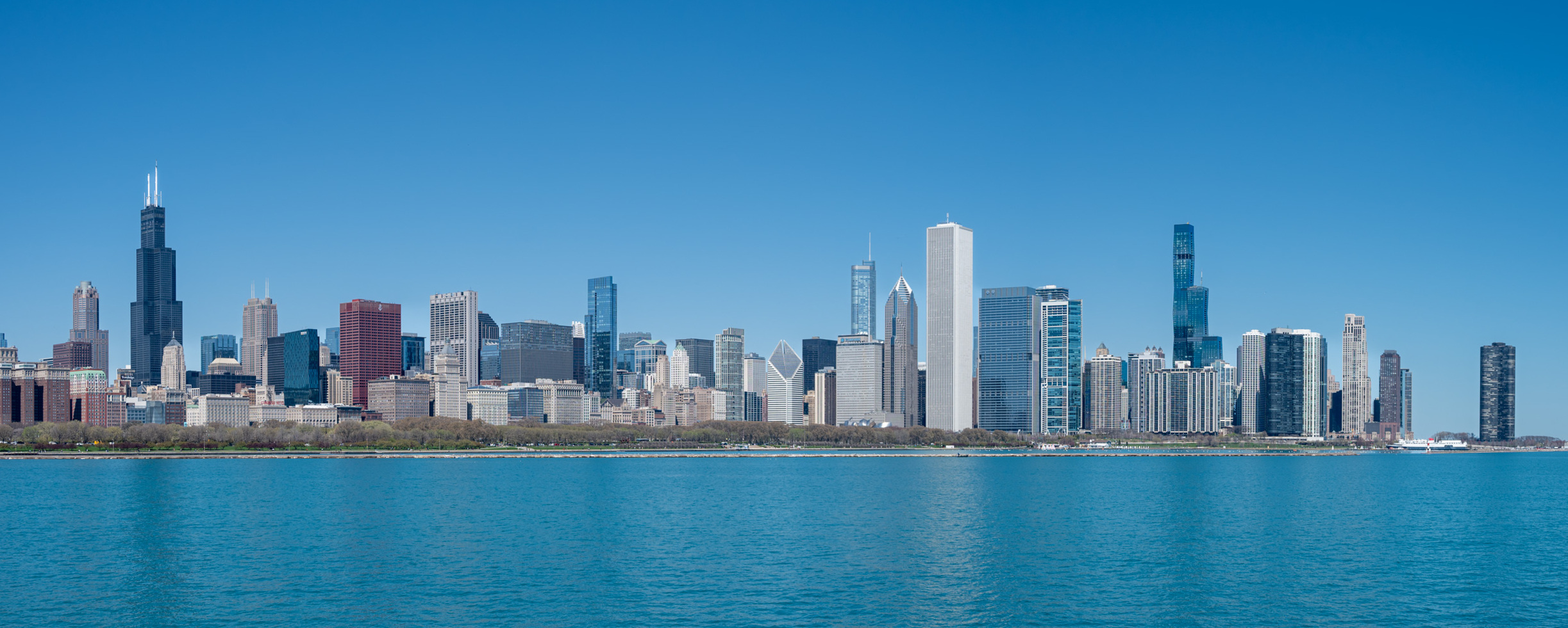Chicago skyscrapers rise above the Lake Michigan shoreline under a clear blue sky.